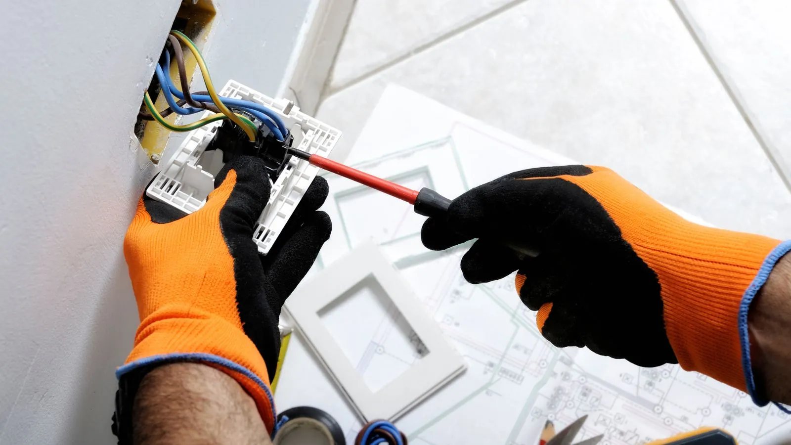 Electrician wiring a wall outlet with blueprints on the floor